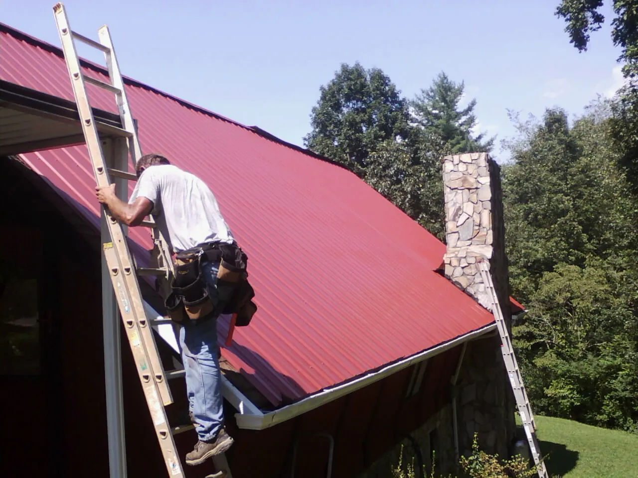Roofer on a ladder beside a red metal roof and stone chimney — Soffit & Fascia Repair by Roofing Brothers in Lynnville